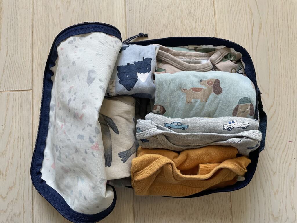 An open packing cube on a wooden floor filled with baby essentials, including patterned onesies and a mustard-colored top, showcasing what to pack for a baby on a plane, ensuring comfort and variety for different needs and temperatures.
