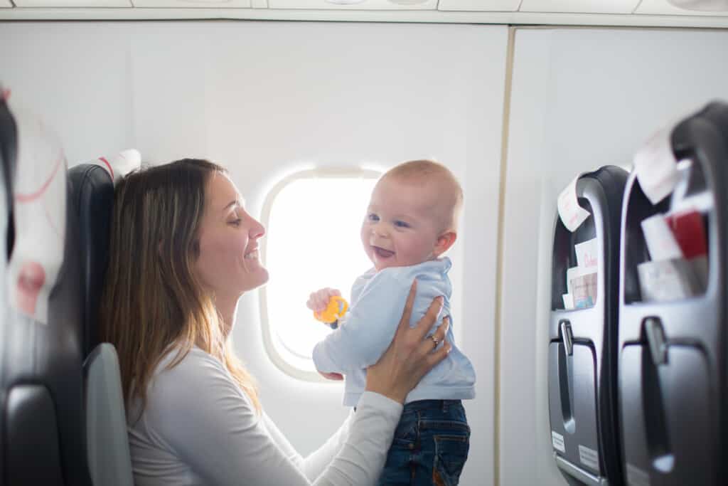 A joyful mother holding her smiling baby aboard an airplane, with a seatback and window in the background, relating to what to pack for baby on plane