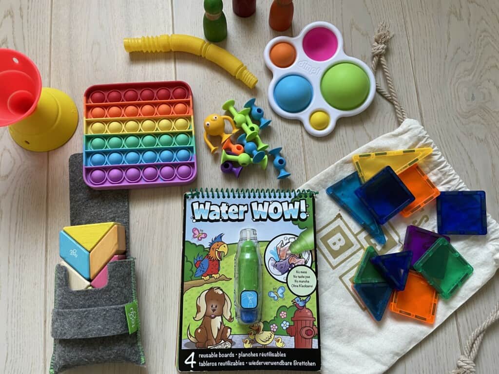 A colorful assortment of the best airplane toys for a 2-year-old, including a pop-it fidget toy, a board book titled 'Water Wow!', and various tactile toys like building blocks, a silicone suction kupp, and transparent magnetic shapes, all spread out on a wooden surface for playtime exploration.