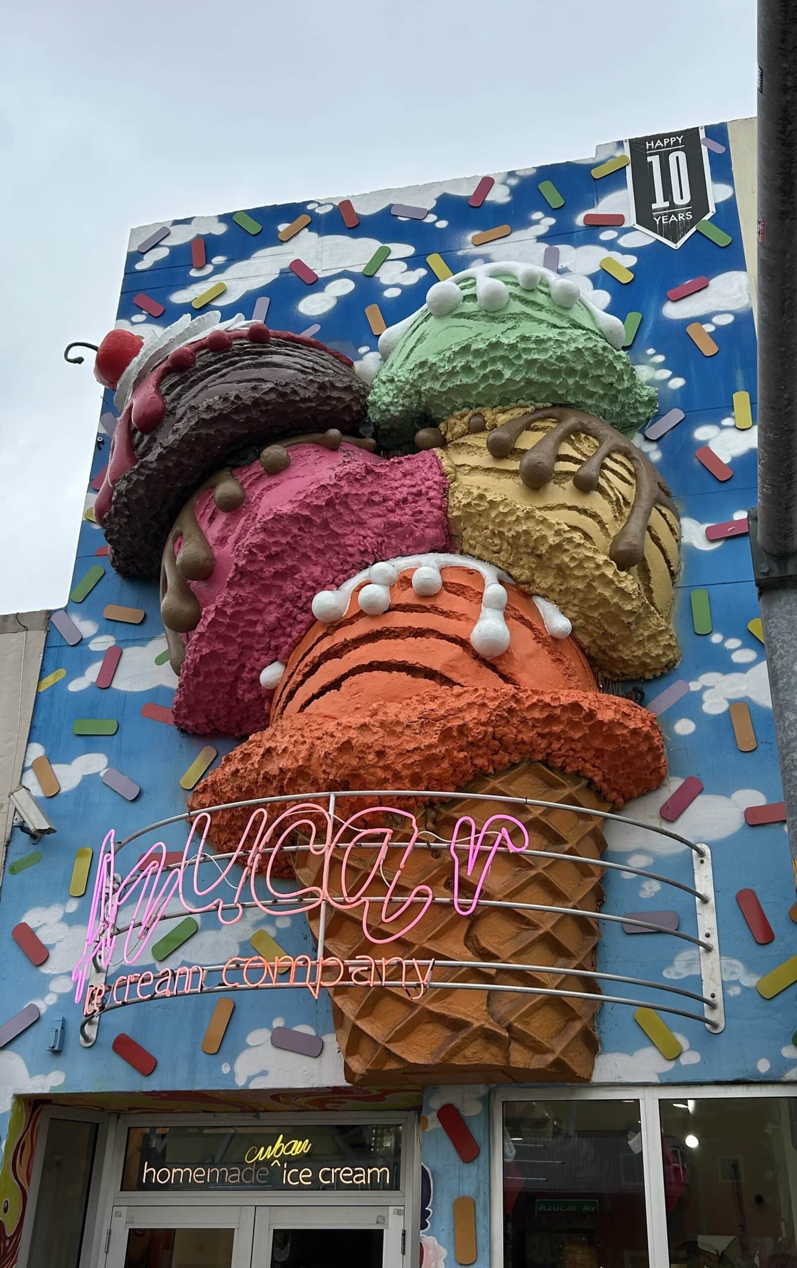 Exterior view of Azucar Ice Cream Company, celebrating 10 years, with a vibrant 3D sculpture of stacked, colorful ice cream scoops in various flavors on top of a waffle cone, against a playful blue wall with scattered sprinkles.