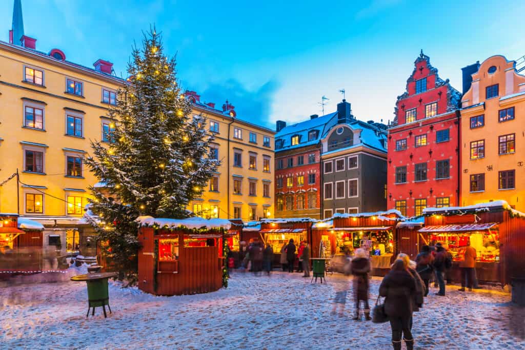 A festive Christmas market in Gamla Stan, Stockholm in winter with kids and families strolling among the red stalls under the glow of a large decorated Christmas tree, capturing the joy and warmth of Swedish winter traditions