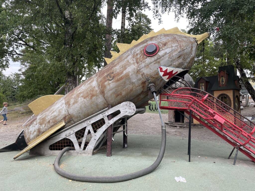 A unique playground feature in Stockholm, showcasing a large, metallic rocket-like structure with a slide, designed to spark imagination, surrounded by greenery and a child playing in the background, highlighting the playful and inventive spirit of Stockholm's play areas.