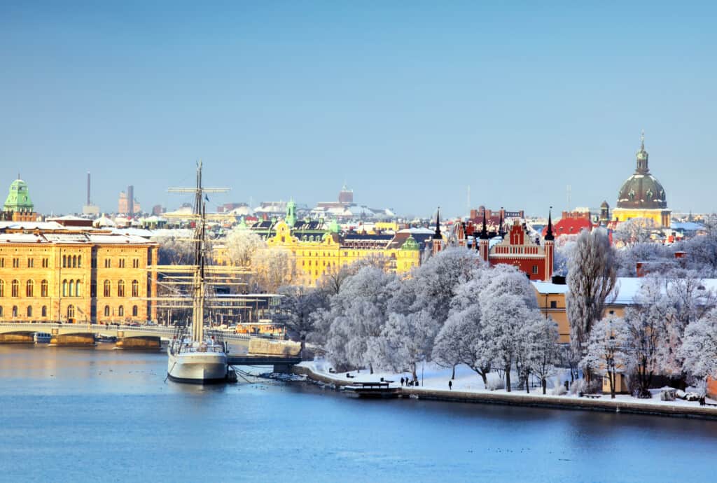A picturesque Stockholm winter panorama featuring frost-covered trees along the waterfront, historic buildings with colorful facades, and a classic tall ship docked in the harbor, inviting exploration and adventure for families visiting Stockholm with kids.
