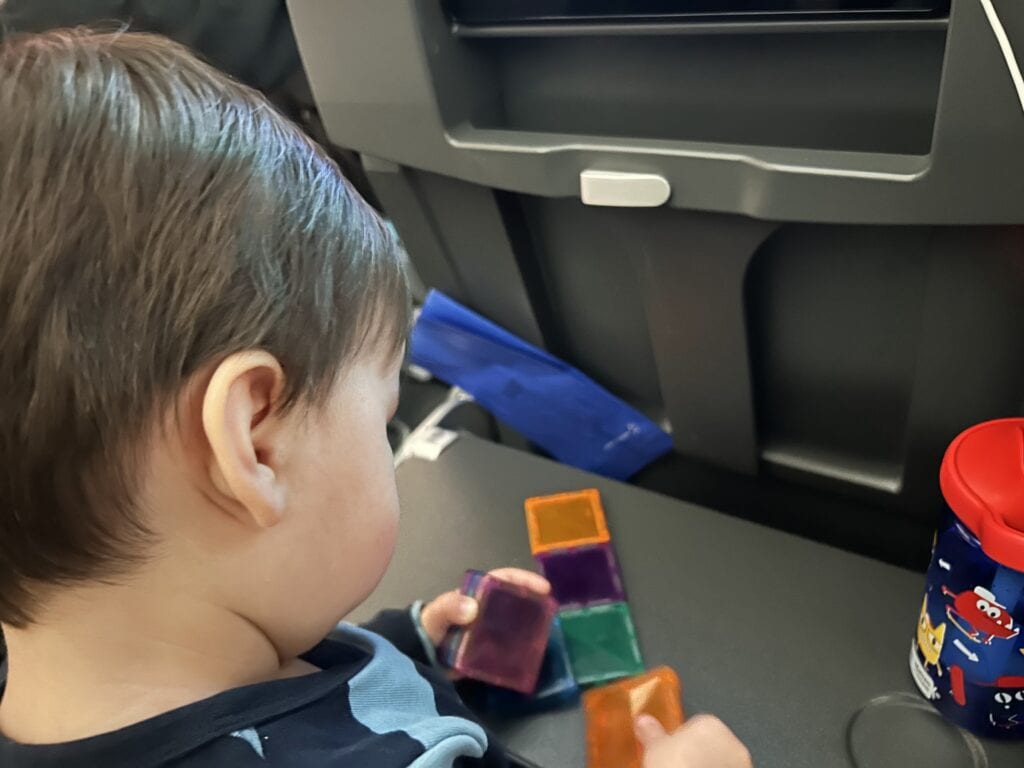 Over-the-shoulder view of a 1-year-old child engaging with colorful magnetic blocks on a fold-down tray in an airplane, showcasing a practical travel toy for a 1-year-old. The child's focus on the toy next to a container of toddler snacks provides a peaceful playtime activity during a flight, emphasizing the importance of portable and entertaining toys for young travelers.