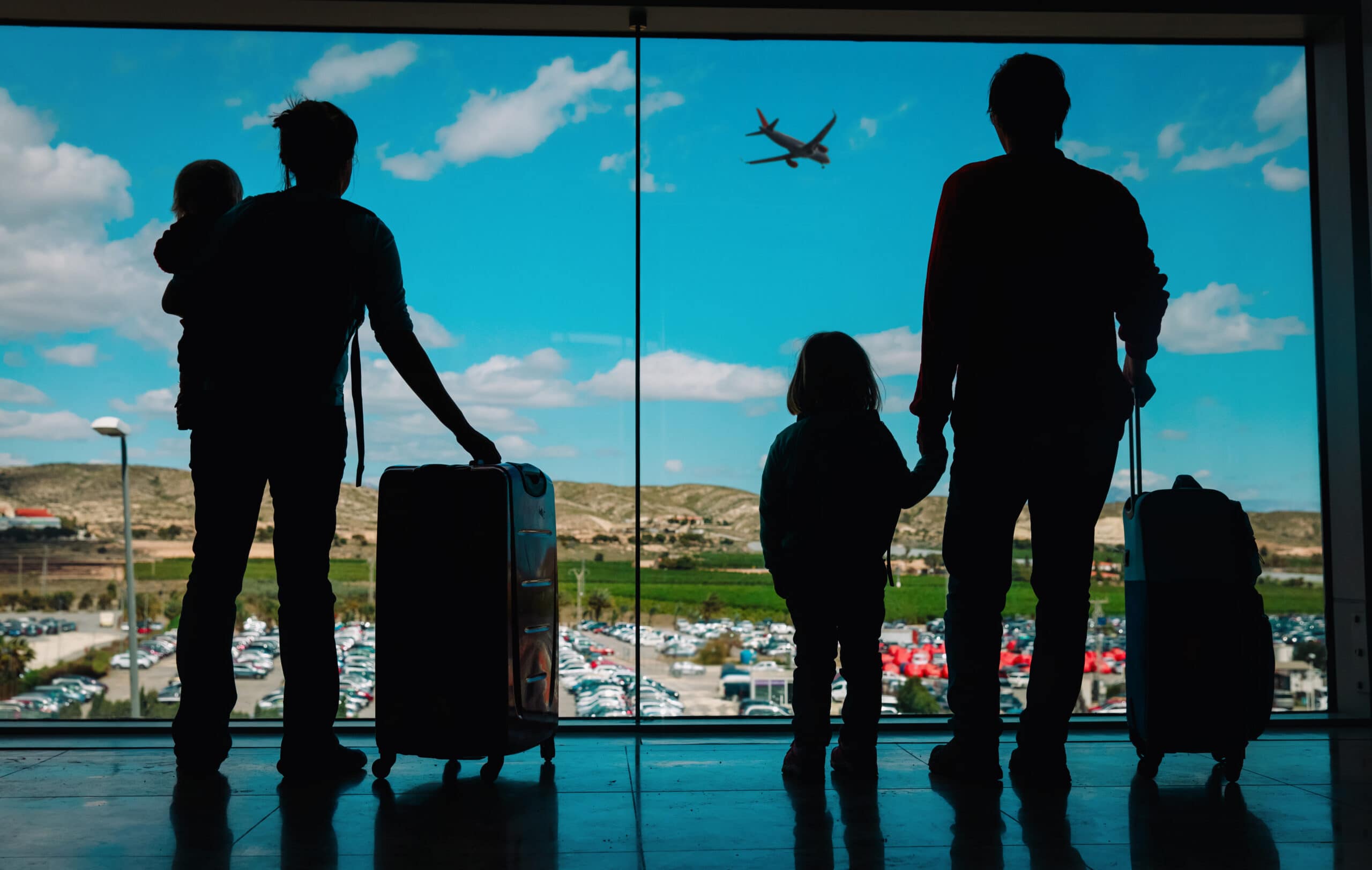 Silhouette of a family at an airport, with two adults and two children looking out a large window at a plane flying in the sky. One adult is holding a toddler, and the other is holding the hand of a small child, with luggage by their sides. This image captures a moment of anticipation and adventure, embodying the spirit of family travel.