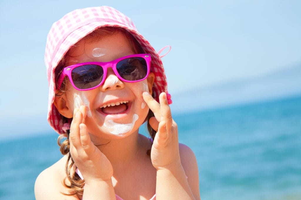 A cheerful young child at the beach, wearing pink sunglasses and a checkered pink hat, with sunscreen applied to her face. She is smiling broadly, representing the joy of sunny beach days and the importance of sun protection, a reminder for travelers to ask, 'Can I pack sunscreen in my checked luggage?' against the backdrop of a clear blue sky and sea.