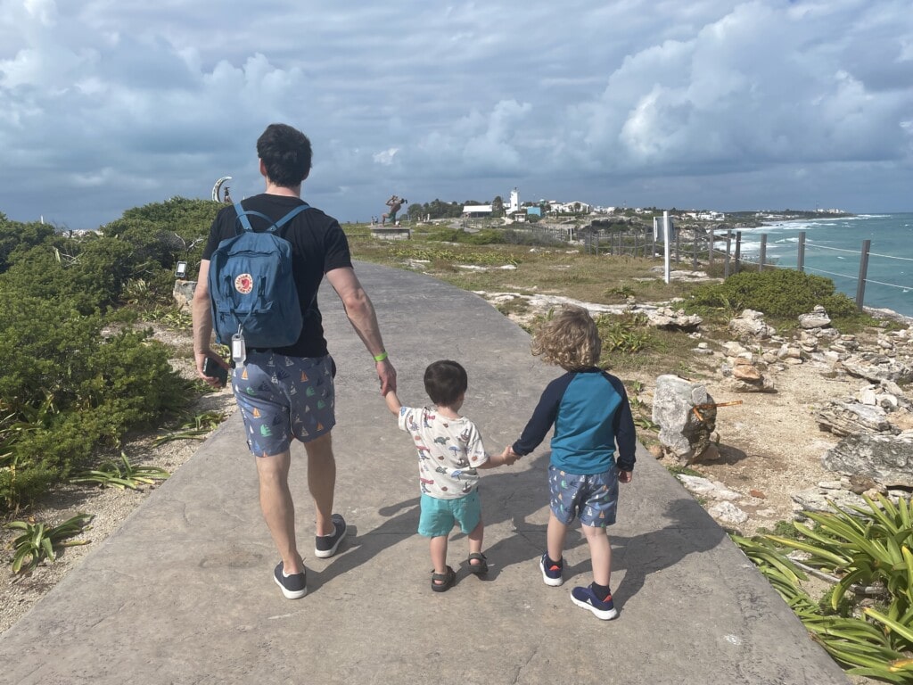 A parent holding hands with two young children walks away from the camera on a curved path by the sea. The stormy sky contrasts with the calm familial scene. The parent is equipped with a minimalist diaper bag, showcasing the bag's sleek design and portability during a family outing near the ocean.