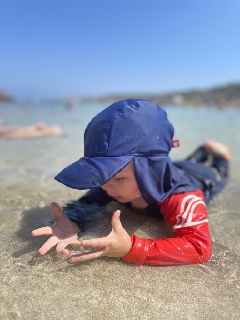A toddler in a red UV protection rashguard and navy hat is captivated by the shallow beach waters, with outstretched hands touching the gentle waves, illustrating the essential sun-safe beachwear for toddlers.