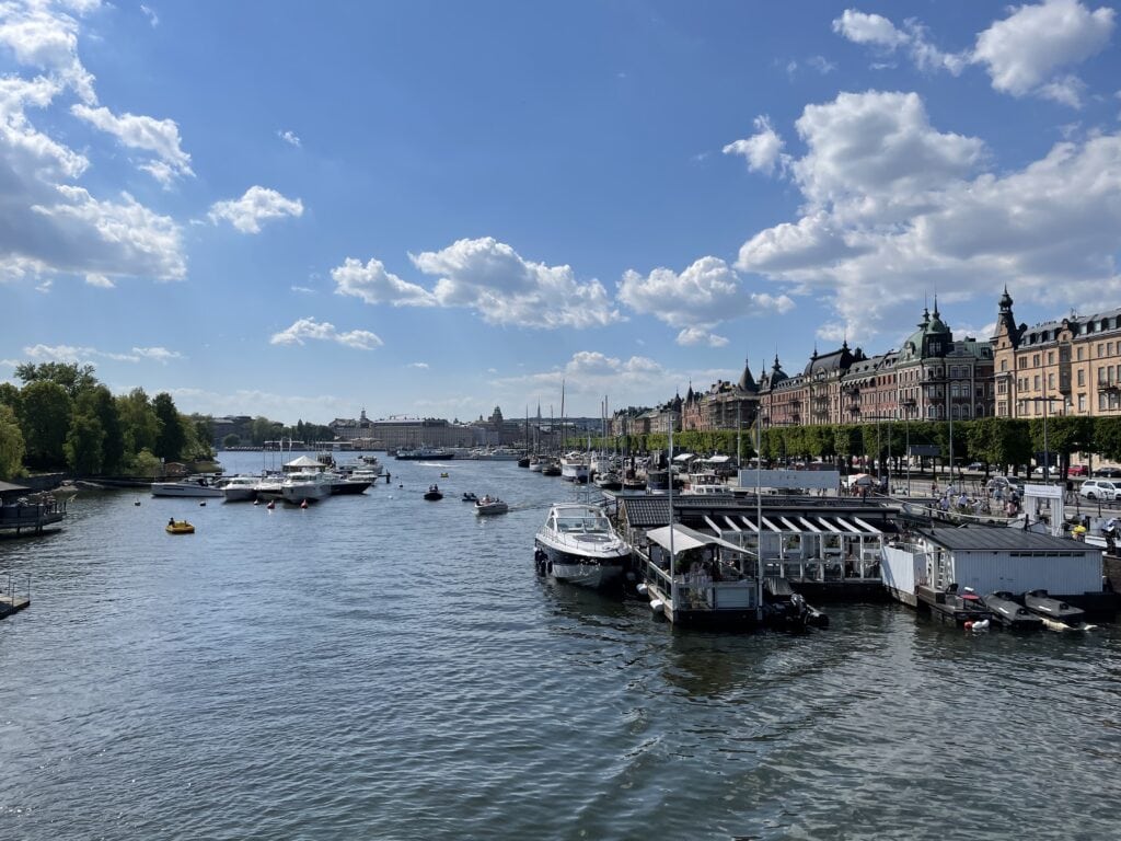 A bustling view of Stockholm's waterfront in the summer, with boats cruising on the water and the city's historic architecture lining the shore. The lively scene captures the essence of a family-friendly atmosphere, perfect for exploring Stockholm with kids during the summer months.