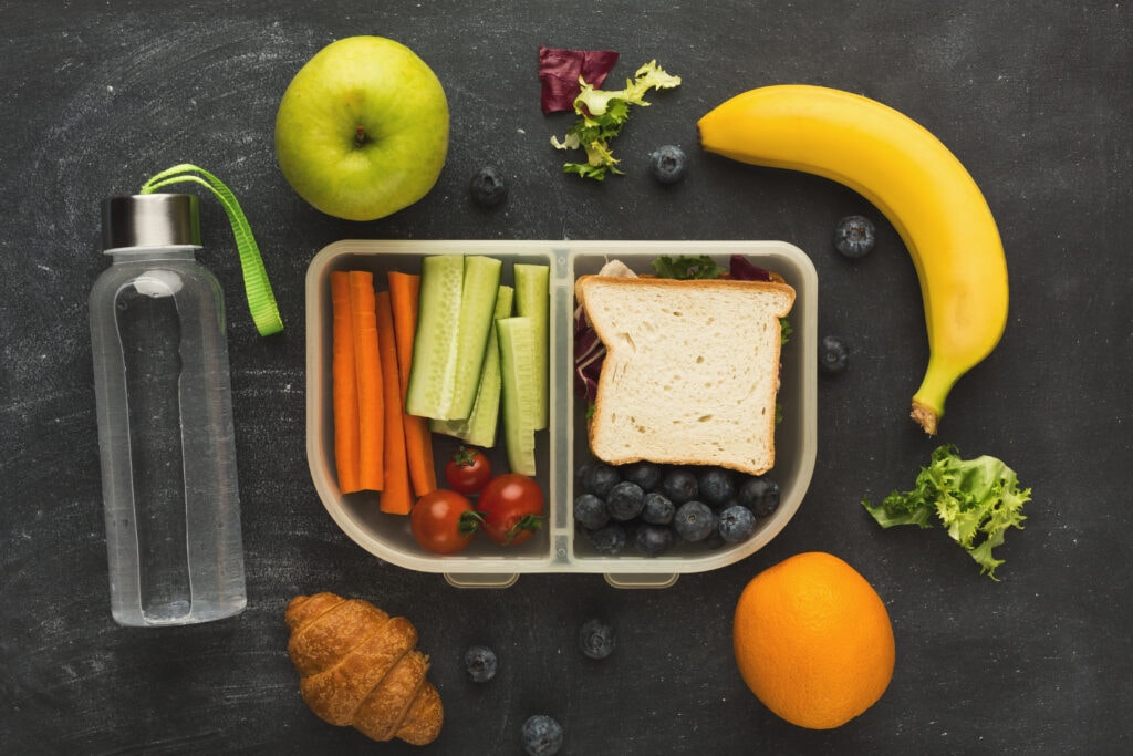 Healthy travel snack ideas for kids displayed on a black background, including a clear water bottle, a green apple, a banana, fresh blueberries, carrot sticks, cucumber slices, cherry tomatoes, a whole grain sandwich, and a croissant. A colorful, nutritious selection for on-the-go eating.