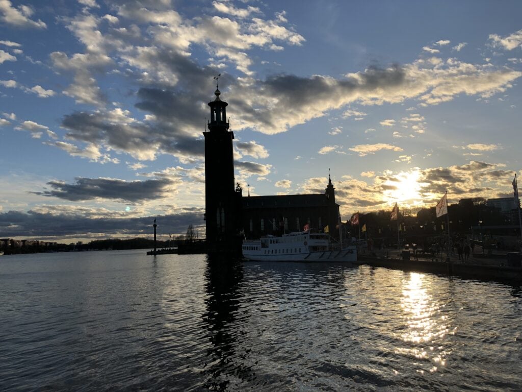 Sunset casting a golden glow over Stockholm's City Hall with its prominent tower silhouetted against a dynamic sky dotted with clouds, viewed from the waterfront, a must-visit landmark to include in a 2 days in Stockholm itinerary.